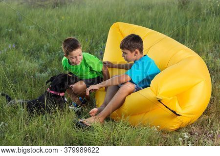 Two Boys Outdoors Sitting On Yellow Air Couch Playing With Dog, Summer Concept