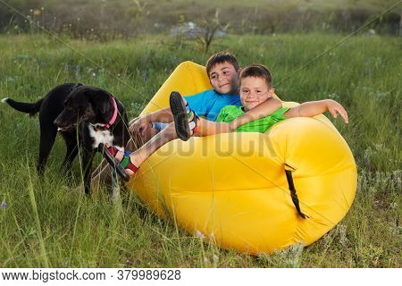 Two Boys In Nature, Lie On A Yellow Lamzak, Next To A Black Dog