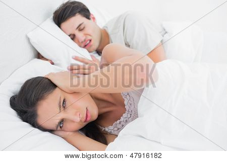 Mujer cubriendo las orejas, mientras que su compañero está roncando a su lado en la cama