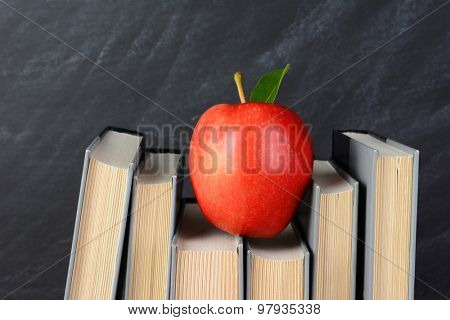 A red apple perched atop text books on a teachers desk with an out of focus chalkboard behind.