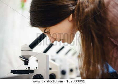 The student girl looks through a microscope in an equipped classroom with a series of microscopes. The concept is to study the science of biology, organisms, bacteria, viruses.