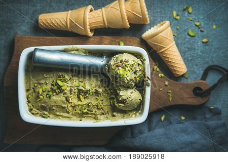 Homemade pistachio ice cream in ceramic mold with metal scooper, crashed pistachio nuts and waffle cones over dark concrete background, top view, selective focus