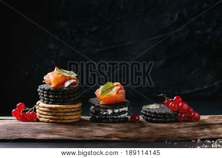 Stack of black charcoal and traditional crackers with smoked salmon, cream cheese, green salad and red currant berries on ood serving board over black metal background. Appetizer snack. Space for text