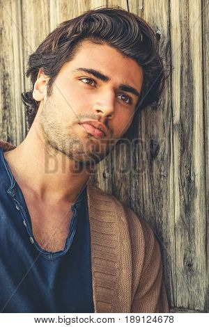 Handsome man relaxing leaning to a wooden wall. The young and attractive man is thoughtful and thinking. Modern hairstyle. Charming casual look. Soft beard.