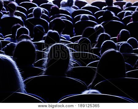 People, children, adults, parents in the theater watching the performance. People in the auditorium looking at the stage. Shooting from the back