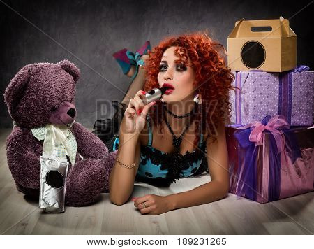 Curly redhead sexy girl looks like a doll for adult men. popsy sucks from a small bottle. Beside sits teddy bear and gift boxes