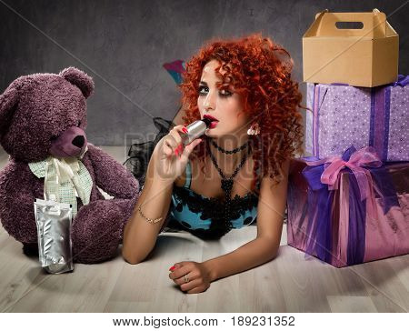 Curly redhead sexy girl looks like a doll for adult men. popsy sucks from a small bottle. Beside sits teddy bear and gift boxes