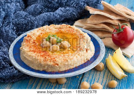 Chickpea paste with garlic, lemon, cumin, sesami seads, olive oil, coriander on a blue ceramic plate on a blue on cloth and wooden background with pita bread lemon and parsley. Healthy eating concept