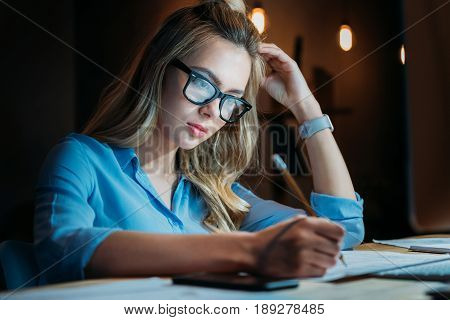 Tired Blonde Caucasian Student In Eyeglasses Writing Something With Pencil And Studying Till Late
