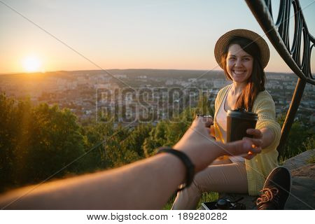 Female giving coffee to man while they sitting on the hill on sunrise city on background