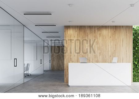 Front view of a white reception desk with two laptops standing on it in front of a wooden office wall. There are glass wall offices to the left. 3d rendering mock up