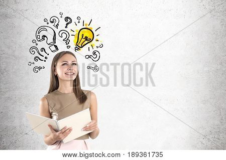 Portrait of a smiling young businesswoman holding a large notebook and standing near a concrete wall with a yellow light bulb surrounded by question marks of different size. Mock up