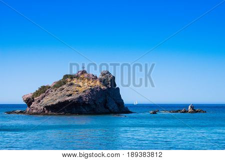 Rock with cross in the blue sea with small sail boat