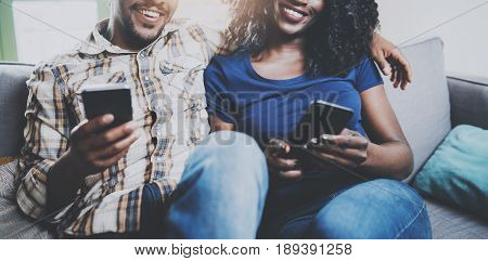 Happy african american couple relaxing together on the sofa.Young black man and his girlfriend using mobile phones while rest at home in the living room.Horizontal, blurred background.Cropped