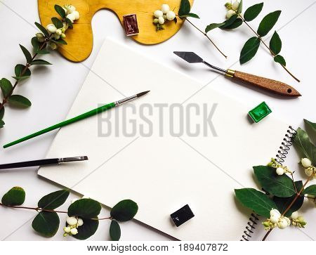 Natural composition with album watercolors and brushes on white background decorated with green snowberry branches and berries. Flat lay top view view from above