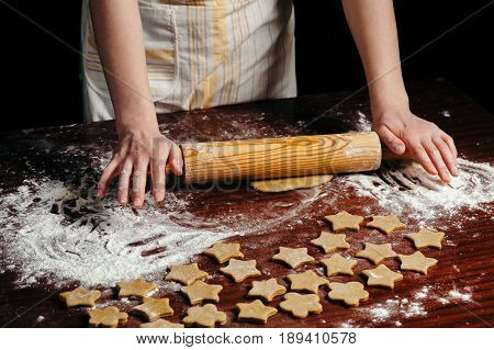 A woman in the kitchen is rolling a dough on a wooden table with a wooden rolling pin. She molds cookies in the form of asterisks.