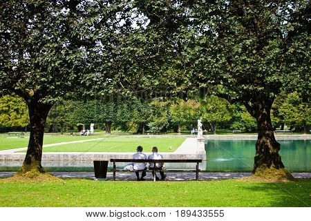 Elderly couple sits on a bench in the park of the Hellbrunn palace. The beautiful park with a pond and trees in which people walk