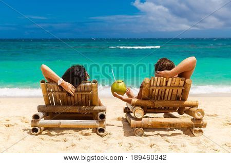 Beach summer couple on island vacation holiday relax in the sun on their deck chairs on the tropical beach. Idyllic travel background.
