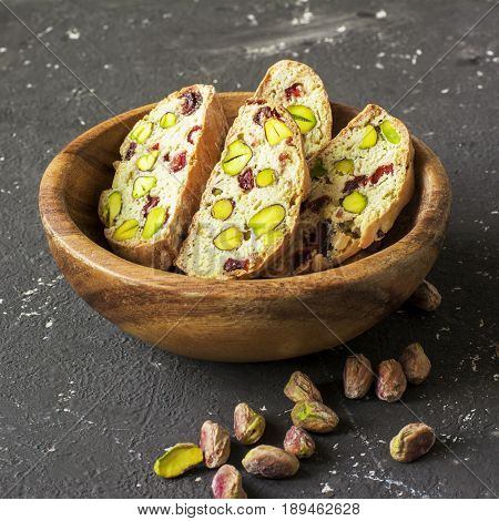 Traditional crispy Italian biscotti or cantuchini crackers with pistachios and dried cranberries in a bowl of olve wood on a dark background. Home classic healthy food