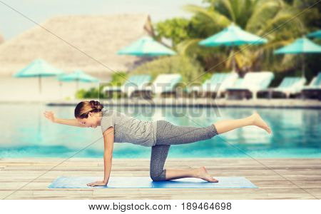 fitness, sport, people and healthy lifestyle concept - woman making yoga in balancing table pose on mat over beach and swimming pool background