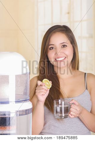 Beautiful smiling woman holding a glass of water behind of a filter system of water purifier on a kitchen background.