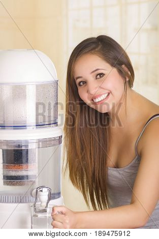Beautiful smiling woman filling a glass of water, with a filter system of water purifier on a kitchen background.