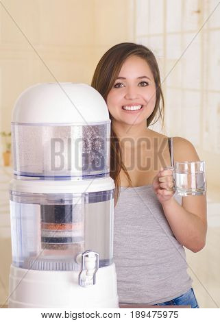 Beautiful smiling woman holding a glass of water behind of a filter system of water purifier on a kitchen background.