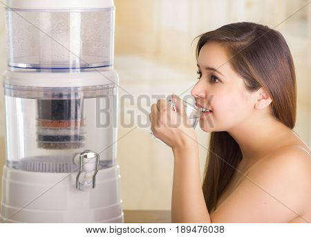 Close up of a beautiful woman drinking a glass of water with a filter system of water purifier on a kitchen background.