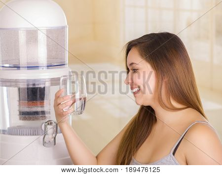 Beautiful smiling woman holding a glass of water, with a filter system of water purifier on a kitchen background.