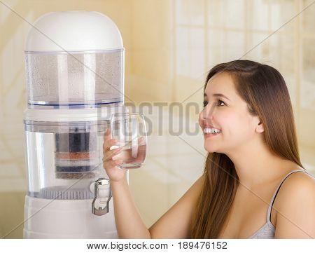 Beautiful smiling woman holding a glass of water, with a filter system of water purifier on a kitchen background.
