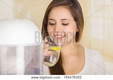 Beautiful woman drinking a glass of water with a filter system of water purifier on a kitchen background.