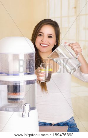 Beautiful woman holding a glass of water in one hand and a pitcher of water in her other hand, with a filter system of water purifier on a kitchen background.