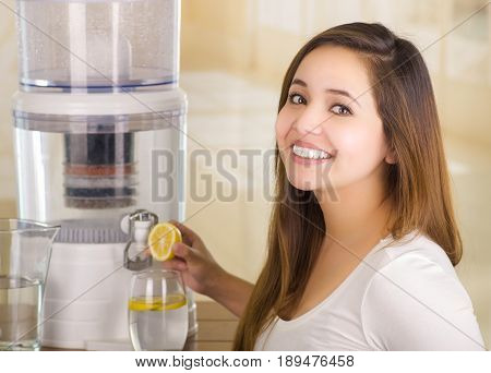 Beautiful smiling woman holding a cut lemon in her hand with a filter system of water purifier on a kitchen background.