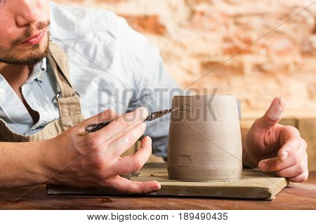 pottery, stoneware, ceramics art concept - closeup on craftsman bent over a clay cup with tool, macro of male hands connecting pieces of fireclay, young male sits at a workshop behind the table