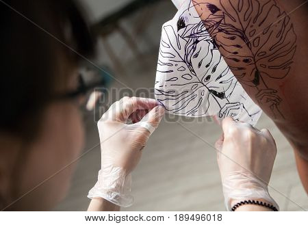 A professional tattooist is presented with sketch on the body of the girl in the Studio. Girl master and model