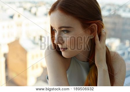 Close up portrait of pretty Caucasian teenage woman looking forward at sunset in big city. Side view of pretty student female touching her colored strands of hair having pensive facial expressions