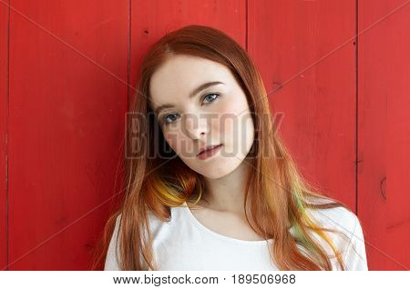 Close up portrait of pretty mixed race teenage girl looking thoughtful and dreamy at camera. Gorgeus redhead student female with colored strands of hair and green eyes standing on red wall background
