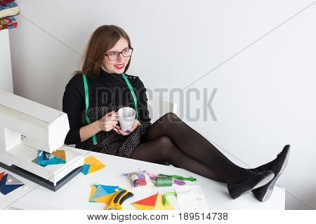 needlework and quilting in the workshop of a tailor - beautiful young woman rests whith a cup, putting her feet up on the table with a sewing machine, fabrics and spools of thread, rotary cutters