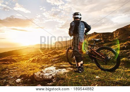 Ryder in full protective equipment on the mtb of two pendant bicycles rides along the green grass towards the sunset in the rays of the sunset sun against the backdrop of a landscape of rocky terrain under the setting sun and clouds.