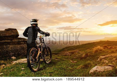 Ryder in full protective equipment on the mtb of two pendant bicycles rides along the green grass towards the sunset in the rays of the sunset sun against the backdrop of a landscape of rocky terrain under the setting sun and clouds.
