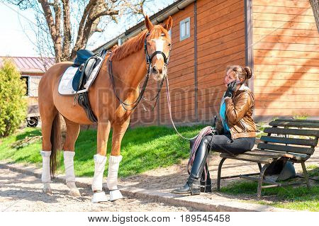 Woman riding trainer near chestnut horse speaking on mobile phone. Horizontal multicolored outdoors image.