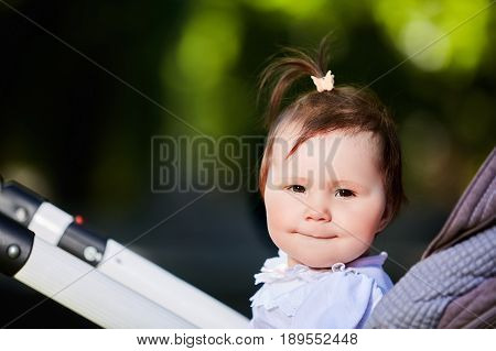 Portrait of the happy baby girl in a stroller in the city park at warm sunshiny day. Cute little girl in the white dress. Summer or spring season in outside. Horizontal photo. Concept of the happy babies.