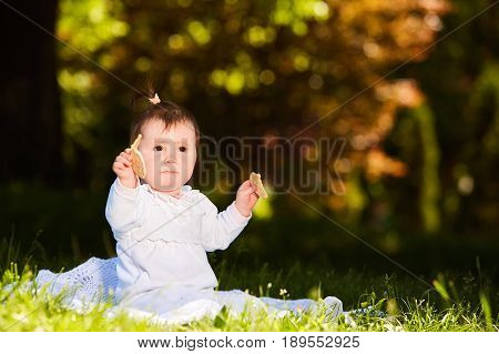 Portrait of happy joyful child sitting on the meadow and holds snack in the park. Cute baby girl in the white dress. Warm sunshiny day. Summer or spring season. Horizontal photo. Concept of the happy babies.