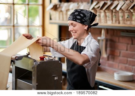 Cheerful female cook rolling pasta knead through machine in cafekithcen. Making pasta. Making pasta in restaurant. Smiling employee cooking pasta in restaurant