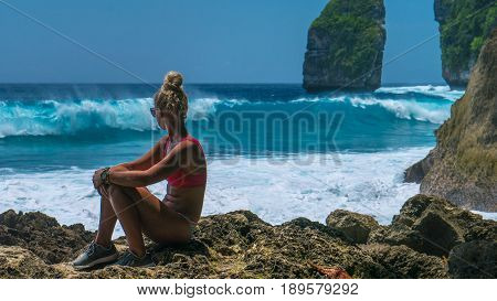 Girl sitting on the Rock and watching Huge Waves hitting Tembeling Coastline at Nusa Penida Island, Bali Indonesia.