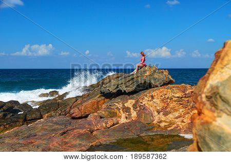 Beautiful young girl seats on big stone rock on rock stone beach on Grete Island Greece. Island rocks beach. Woman in red. Greece islands holidays vacations tours famous sightseeing destinations