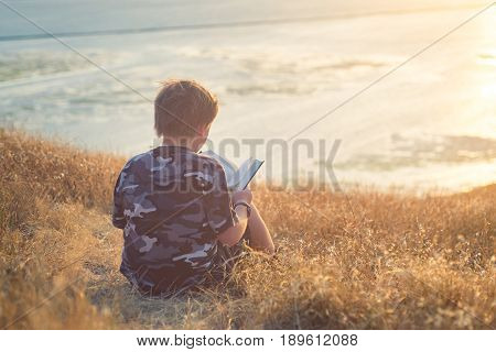 Boy reading book in yellow grass high in the hills, watching beautiful sunset above Bay Area, rear view