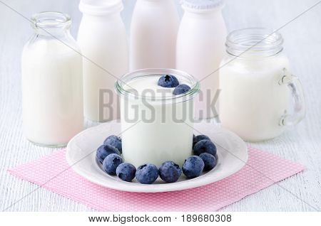 a cup of yogurt with blueberrys and different bottles of drink yogurt and milk on a plate, napkin on the table