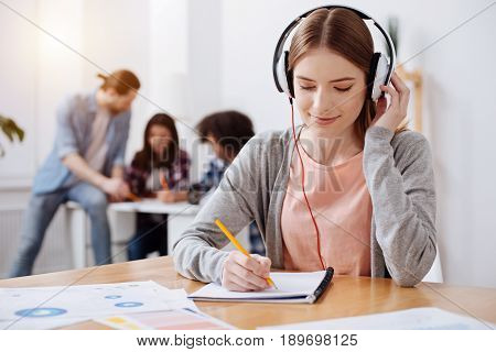 Selecting important pieces. Beautiful smart motivated lady wearing headphone and listening to audio lesson while making notes in her notebook