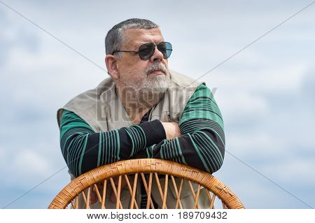 Outdoor portrait of senior man in black sunglasses leant on a wicker chair back and looking